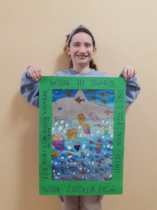 Uczennica Maria z klasy 6a trzyma swoją pracę plastyczną. Na zielonym marginesie widnieje napis Woda to skarb, dbaj o rzeki, morza, oceny. Praca przedstawia ryby, meduzy oraz pływającego w wodzie wieloryba. pływającego w morzu.
